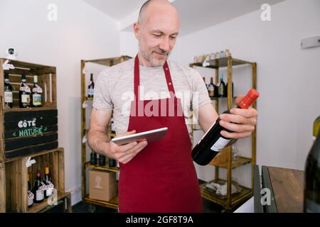 Reifer Verkäufer Mann mit einer Tablette, während eine Flasche Wein in einem Weinladen halten Stockfoto