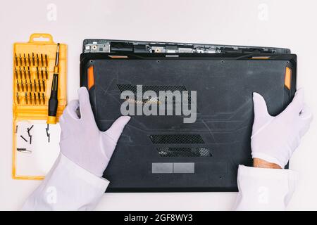 Betrachten Sie von oben einen mit weißen Handschuhen bedeckten Techniker, indem Sie den Deckel eines Laptops entfernen, bevor er auf einem weißen Tisch repariert wird Stockfoto