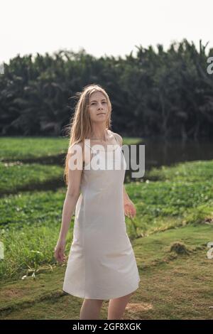 Porträt einer hübschen kaukasischen Frau in weißem Kleid, die im Park spazierend und lächelnd unterwegs ist. Lange blonde Haare. Natürliche Schönheit. Make-up frei. Schöner Sonnenschein Stockfoto