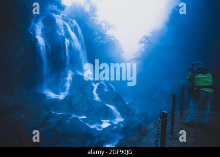 Touristenmann, der mit der Kamera vor einem Wasserfall in Nebel fotografiert. Touristische Attraktionen in Sa Pa, Vietnam. Langzeitbelichtung. Stockfoto