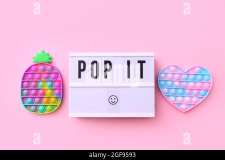 Trendy beliebtes Silikon bunte Anti Stress Pop IT Spielzeug für Kinder auf rosa Hintergrund mit Text Pop IT auf Leuchtkasten. Stockfoto