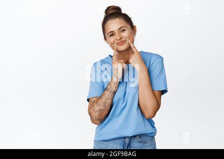 Nette junge Frau lächelnd, zeigt ihre Grübchen auf den Wangen und steht hübsch in blauem T-Shirt vor weißem Hintergrund Stockfoto