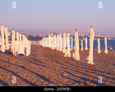 Gefaltete Sonnenschirme und Sonnenliegen stehen im Abendlicht auf dem Kiesstrand Stockfoto