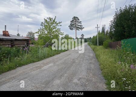 Nahaufnahme einer Uferstraße aus kleinen Steinen, grünes hohes Gras wächst entlang der Straße, und in der Ferne gibt es kleine Holzhäuser. Hochwertige Fotos Stockfoto