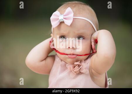 Das lustige Mädchen nahm ein rotes Band in den Mund. Ein Jahr altes Kind. Ein niedliches Baby. Stockfoto