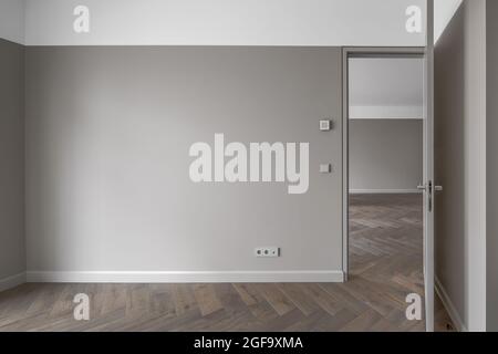 Einfache, moderne beigefarbene Wand mit grauem Lichtschalter und grauer Steckdose im Empty Room mit offener Tür und Eichenholzboden. Interior Design Element von c Stockfoto