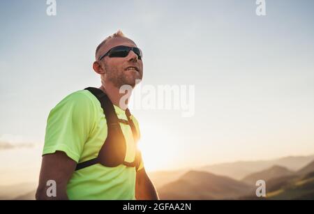 Portrait von Active Mountain Trail Läufer gekleidet helles T-Shirt mit Rucksack und Sport Sonnenbrille ruhen, während er Himmel läuft durch malerische Hügel ein Stockfoto