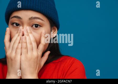 Schockiert verängstigte junge asiatische Frau in legerer Kleidung, die über blauem Wandhintergrund steht, bedeckt das Gesicht mit den Händen, Nahaufnahme Stockfoto