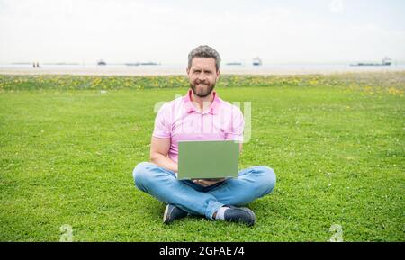 Glücklicher Geschäftsmann mit Laptop zum Bloggen auf grünem Gras, Freiberufler Stockfoto