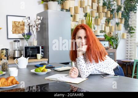 Lächelnde attraktive Frau mit roten Haaren sitzt mit Notizblock und Stift in der Küche zu Hause. Mutterschaftsunterricht, Fernunterricht und Home Office Stockfoto