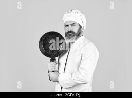 Antihaftbeschichtetes Kochgeschirr. Braten ohne Öl. Professionelle Küchenutensilien. Bärtiger Koch bereitet das Frühstück zu. Antihaftpfanne zum Braten. Emaillierte Kochgefäße Stockfoto