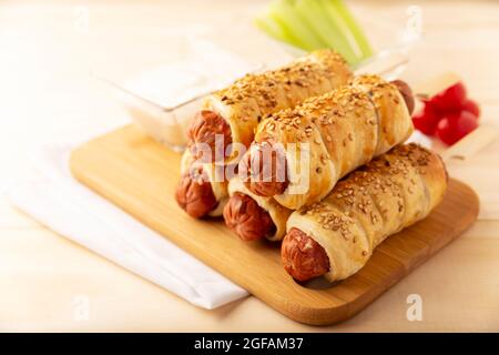 Köstlicher Snack mit Würstchen, eingewickelt in Blätterteig mit Sesam, begleitet von Kirschtomaten, Sellerie und Sauce. Serviert auf Holzbrett Stockfoto