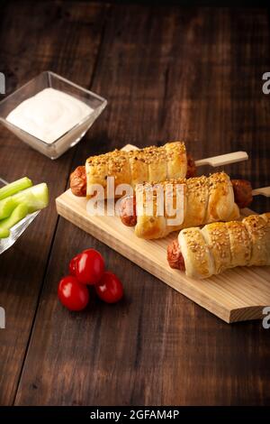 Köstlicher Snack mit Würstchen, eingewickelt in Blätterteig mit Sesam, begleitet von Kirschtomaten, Sellerie-Sticks und Sauce. Serviert auf Holzbrett Stockfoto