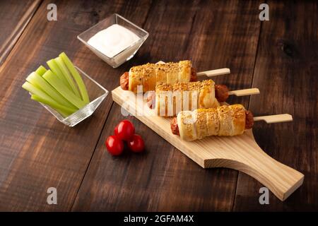 Köstlicher Snack mit Würstchen, eingewickelt in Blätterteig mit Sesam, begleitet von Kirschtomaten, Sellerie-Sticks und Sauce. Serviert auf Holzbrett Stockfoto