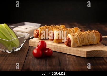 Köstlicher Snack mit Würstchen, eingewickelt in Blätterteig mit Sesam, begleitet von Kirschtomaten, Sellerie-Sticks und Sauce. Serviert auf Holzbrett Stockfoto