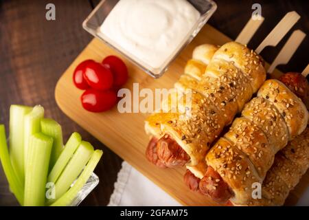 Nahaufnahme eines leckeren Snacks mit Würstchen, eingewickelt in Blätterteig mit Sesam, begleitet von Kirschtomaten, Sellerie-Sticks und Sauce. Serviert Stockfoto