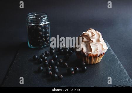 Appetitlich gebackener Vanillemuffin mit Johannisbeeren auf einem schwarzen Servierbrett. Köstlicher Cupcake aus Schokolade auf dunklem Hintergrund mit verstreuten Johannisbeeren Stockfoto