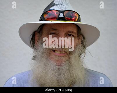 Bärtig optimistischer älterer kaukasischer Mann lächelt und trägt einen bemalten Panama-Hut mit farbenfroher Sonnenbrille am Rand seines Hutes, weißer Hintergrund. Stockfoto