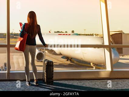 Reisen Sie während der Coronavirus-Pandemie-Touristen mit Gepäck und beobachten Sie den Sonnenuntergang am Flughafenfenster. Nicht erkennbare Frau, die in der Lounge am Boarding Gate wartet Stockfoto