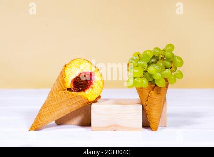 Eine halbe reife Pfirsich- und grüne Traube in süßem Waffelkegel auf Holz steht auf hellorangefarbenem Hintergrund. Gesunde Ernährung, saisonale Ernte der Früchte. Co Stockfoto