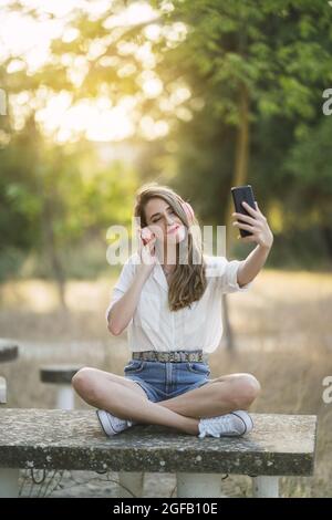 Junge Spanierin trägt Shorts mit einem Hemd und nimmt ein Selfie mit einem Telefon in einem Park Stockfoto