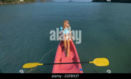 Die ziemlich schlanke Frau im eleganten Badeanzug liegt im rosa Plastikkajak, das auf dem azurblauen Ozean in der exotischen Urlaubsoberansicht driftet. Stockfoto