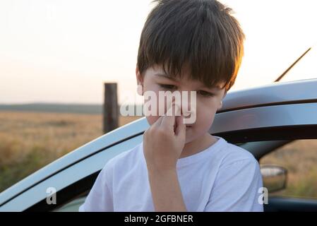 Junge, der bei Sonnenuntergang neben einem Auto seine Nase pflückt. Trockenes und heißes Wetter bringt Unwohlsein und Atembeschwerden mit sich. Stockfoto