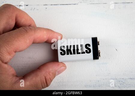 KOMPETENZKONZEPT. Weiße Batterie in der Hand eines Mannes auf einem bemalten Holzhintergrund. Stockfoto