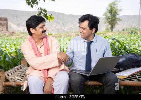 Glücklich lächelnd ländlichen indischen männlichen Landwirt Händeschütteln mit Agronom oder Bankier verwenden Laptop auf dem Bett auf dem Feld Landwirtschaft sitzen. Angebot endgültig, finanzielle unterstützung Stockfoto