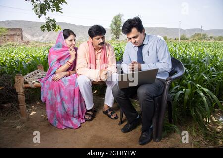 Glückliche ländliche indische Bauernfamilie mit Agronom oder Bankier verwenden Laptop. Ein Beamter, der die Richtlinie auf dem Computerbildschirm, der finanziellen Unterstützung oder der cro für die Landwirtschaft zeigt Stockfoto