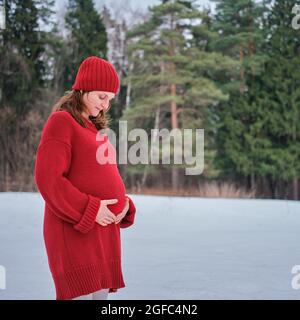 Eine glückliche Schwangere in einem Strickpullover und einem Hut spaziert im Park, einem Winterwald mit Bäumen im Schnee Stockfoto