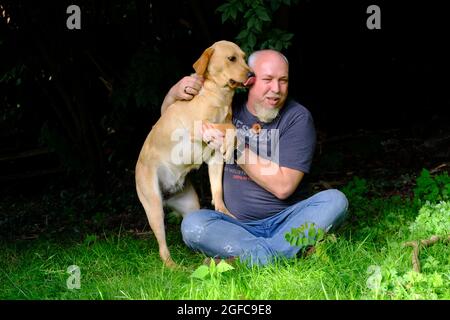 Schöner Mann spielt mit seinem glücklichen goldenen labrador Retriever Hund auf dem Garten Rasen. Der Mann hat Spaß mit dem loyalen Stammhund im Freien im Sommerhaus-Garten. Hochwertige Fotos Stockfoto