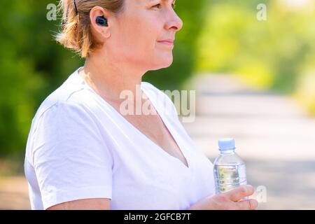 Das Profilfoto einer sportlich blonden erwachsenen Frau, die Ohrenhilfe trägt und die Flasche Wasser in den Händen hält und sich nach dem Joggen im Freien ausruht. Stockfoto