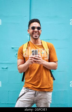 Fröhlicher junger Latino-Mann, der die Kamera anschaut, während der Rucksack auf seinem Handy tippt und die Straße entlang läuft, trägt ein orangefarbenes T-Shirt mit lgtbi-Aufkleber und einer Sonnenbrille. Hochwertige Fotos Stockfoto