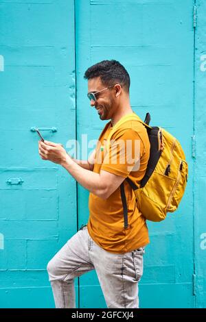 Fröhlicher junger lateiner mit seinem Rucksack, der auf seinem Handy tippt, der die Straße entlang geht und ein orangefarbenes T-Shirt und eine Sonnenbrille trägt. Hochwertige Fotos Stockfoto