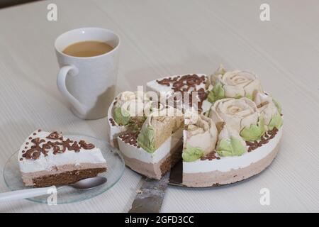 Vogelmilchkuchen, verziert mit marmelgelbroten Rosen und Schokolade. In Stücke schneiden. In der Nähe befindet sich eine Tasse Kaffee mit Milch. Stockfoto