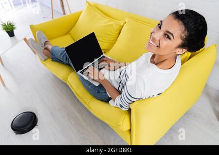 Aus dem hohen Winkel betrachtet, wie ein freiberufler aus dem afrikanischen amrican mit einem Laptop auf der Couch in der Nähe des Roboter-Staubsaugers arbeitet Stockfoto