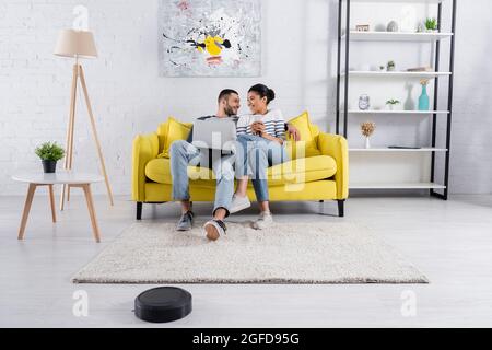 Lächelndes, multiethnisches Paar, das zu Hause Geräte in der Nähe eines Roboter-Staubsaugers verwendet Stockfoto