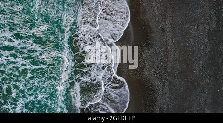 Seeufer mit dramatischer Textur aus brechenden Wellen und schwarzem Sand, Drohnenperspektive Stockfoto