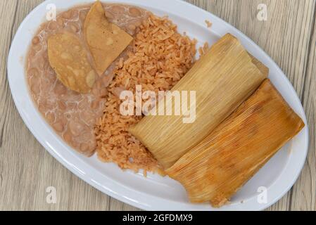 Blick von oben auf einen herzhaften Teller mit Tamales, eingewickelt in Maisschalen, serviert mit Reis und gebratenen Bohnen. Stockfoto