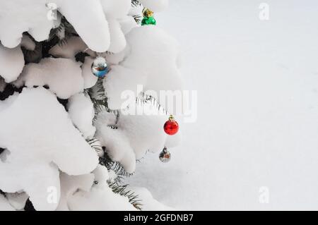 Eine Nahaufnahme eines Weihnachtsbaums im Freien mit stark schneebedeckten Ästen, die mit blauen, roten und grünen Weihnachtskugeln verziert sind Stockfoto
