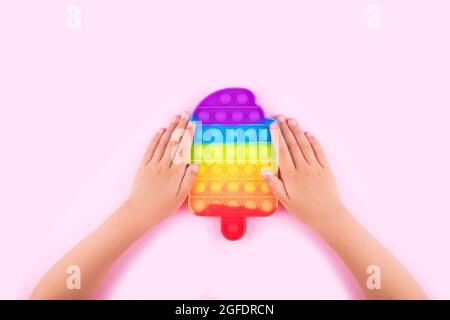 Kinderhände halten Regenbogen Pop it Anti-Stress-Spielzeug isoliert auf rosa Hintergrund. Nahaufnahme. Stockfoto