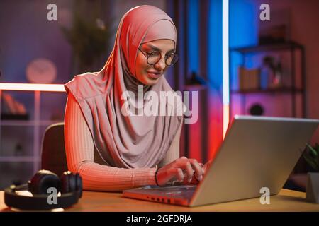 Attraktive, selbstbewusste junge, qualifizierte arabische Frau im Hijab, die zu Hause an ihrem entfernten Arbeitsplatz sitzt und auf dem Laptop tippt. Bunte Nachtlichter auf dem Hintergrund Stockfoto