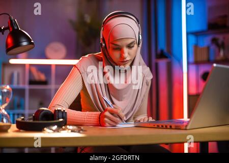 Eine sympathisch fokussierte, selbstbewusste 25-jährige araberin mit Kopftuch und Headset, die sich Notizen macht, während sie Online-Webinare oder -Lektionen auf dem Laptop hört. E-Learning zu Hause Konzept. Fernstudium Stockfoto