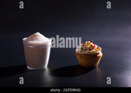 Hoher Zuckergehalt in verschiedenen Desserts. Glas Zucker mit Tasse-Kuchen, Nahaufnahme auf schwarzem Hintergrund. Stockfoto