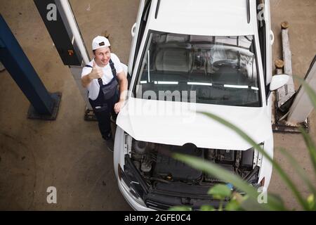 Draufsicht auf einen Mechaniker, der mit offener Haube zur Kamera in der Nähe des Autos lächelt Stockfoto