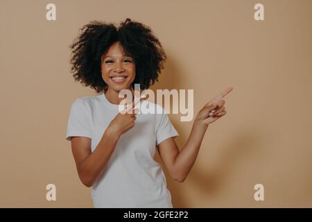 Positive, fröhliche, dunkelhäutige Frau, die auf einen auf beigem Hintergrund isolierten Kopierraum zeigt Stockfoto