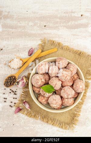 Gefrorene Fleischbällchen mit Gewürzen, Basilikumblättern und Knoblauchzehen in einer Keramikschale. Rohe Zutaten zum Kochen von Lebensmitteln. Holzhintergrund, Draufsicht Stockfoto