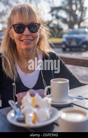Ältere Erwachsene Frau, die lächelt und eine Brille mit Latte und Zitronenkuchen trägt. Blick auf die Kamera Stockfoto