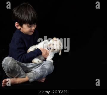 Barfuß brünett kleiner Junge sitzt auf dem Boden und spielt mit seinem Pudel Welpen im Studio, leichte Aprikose. Horizontal, schwarzer Hintergrund, Kopierraum Stockfoto